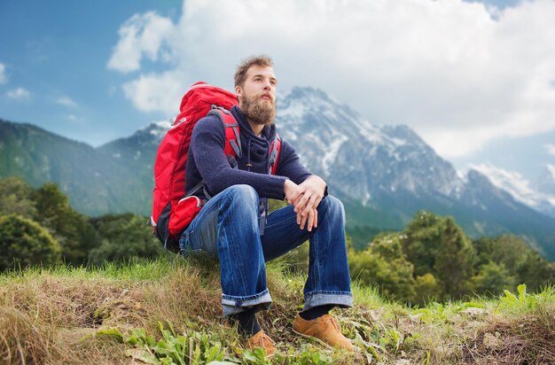 Abenteuer-, Reise-, Tourismus-, Wander- und Personenkonzept - lächelnder Mann mit rotem Rucksack, der auf dem Boden über alpinem Berghintergrund sitzt