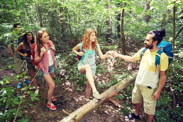 Abenteuer-, Reise-, Tourismus-, Wanderungs- und Personenkonzept - eine Gruppe lächelnder Freunde, die mit Rucksäcken spazieren gehen und über umgestürzte Baumstämme im Wald klettern