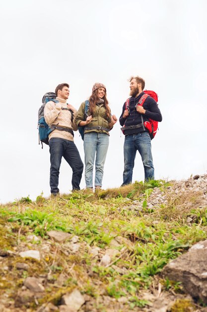 Abenteuer-, Reise-, Tourismus-, Wanderungs- und Personenkonzept - eine Gruppe lächelnder Freunde mit Rucksäcken, die auf einem Hügel stehen und sich im Freien unterhalten