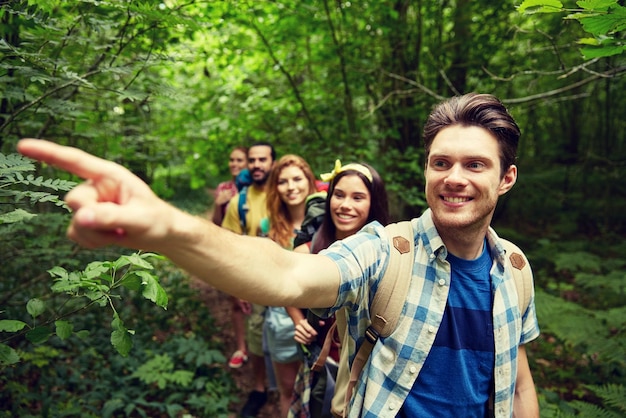 Abenteuer-, Reise-, Tourismus-, Wanderungs- und Personenkonzept - Gruppe lächelnder Freunde mit Rucksäcken, die mit dem Finger in den Wald zeigen