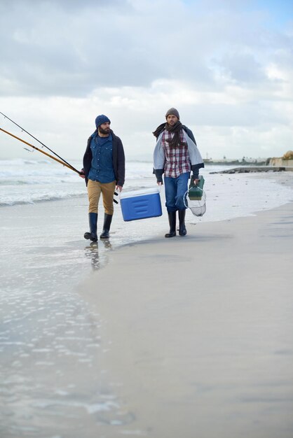 Foto abenteuerfischen und männer, die zusammen mit kühlgeräten auf dem strand spazieren gehen, und urlaubsverbindungen meeresfischer und freunde mit stangen, köder und werkzeugen bei wellen am wintermorgen, urlaub auf see