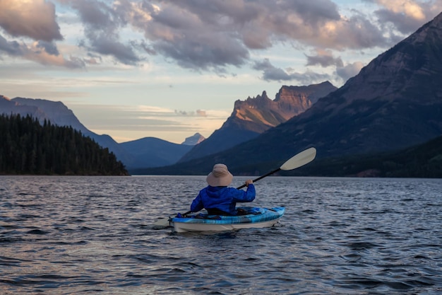 Abenteuerlustiger Mann beim Kajakfahren im Glacier Lake, umgeben von den wunderschönen kanadischen Rocky Mountains, während eines bewölkten Sommersonnenuntergangs, aufgenommen im Upper Waterton Lake, Alberta, Kanada