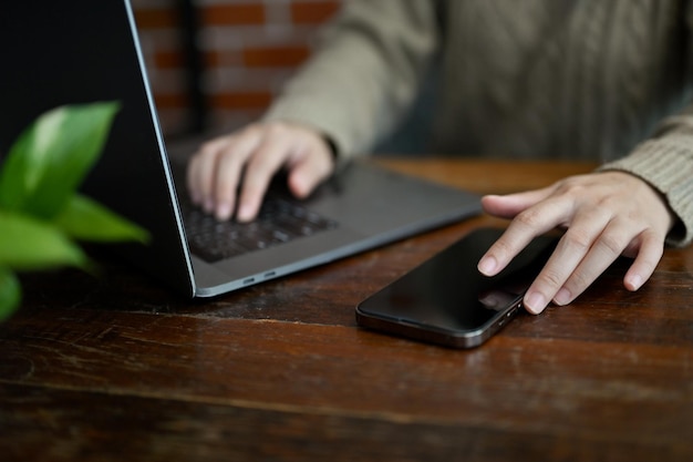 Abgeschnitten Eine Frau, die an ihrem Laptop arbeitet, während sie ihr Smartphone am Tisch benutzt