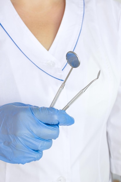 Abgeschnittene Ansicht des Zahnarztes in Latexhandschuhen, die das professionelle Werkzeug des Zahnarztes in der Nähe des Zahnsets halten, das auf dem einheitlichen Hintergrund des Arztmantels verschwommen ist
