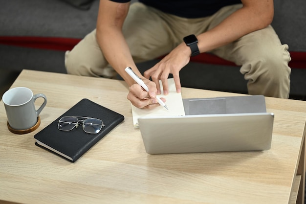 Abgeschnittene Ansicht eines Mannes, der einen Laptop benutzt und sich Notizen auf dem Notizblock macht, während er zu Hause aus der Ferne arbeitet