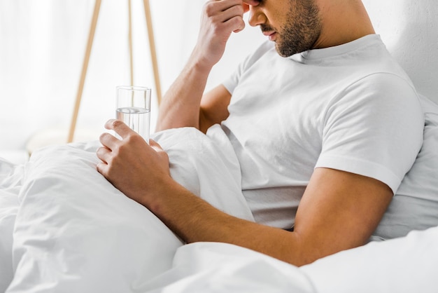 Abgeschnittene Ansicht eines Mannes mit Kopfschmerzen, der ein Glas Wasser im Bett hält