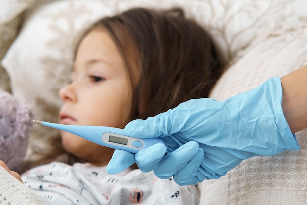 Abgeschnittene Arzthand im Handschuh halten elektronisches Thermometer gegen ruhiges trauriges stilles kleines Mädchen im Bett Quarantänezeit