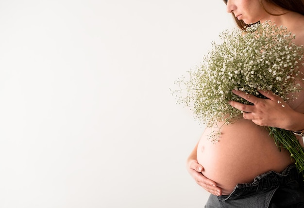 Abgeschnittene Aufnahme einer nicht erkennbaren nackten schwangeren Frau, die die Brust mit einem großen Gypsophila-Blumenstrauß versteckt, der den Bauch auf weißem Hintergrund berührt und streicheltSchöne Schwangerschaft im FrühlingStillen