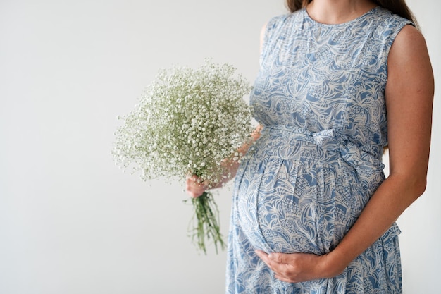 Abgeschnittene Aufnahme einer nicht erkennbaren schwangeren Frau mit Blumen, die den Bauch berührt und streichelt, auf weißem Hintergrund. Schöne, gesunde, glückliche Schwangerschaft im Frühlingssommer