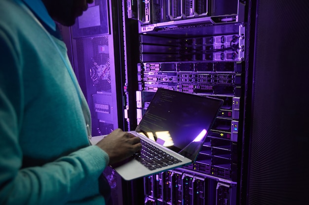 Abgeschnittene Aufnahme eines afroamerikanischen Dateningenieurs, der einen Laptop hält, während er mit einem Supercomputer im Serverraum arbeitet, der von blauem Licht beleuchtet wird, Kopierraum