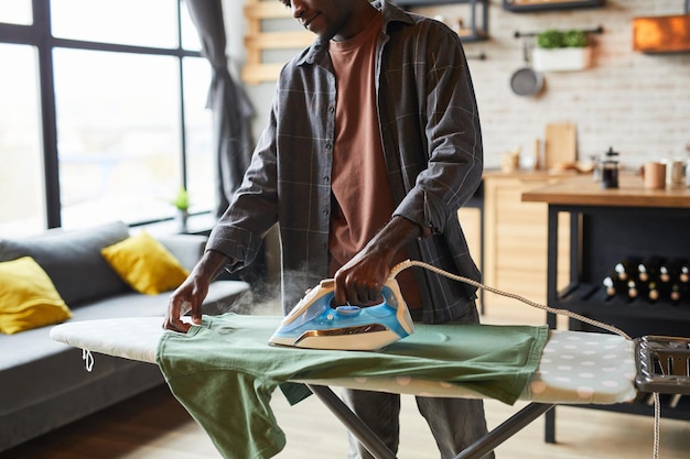 Abgeschnittene Aufnahme eines jungen afroamerikanischen Mannes, der Kleidung in einer Einzimmerwohnung bügelt, Junggesellenlebensstil und Konzeptkopienraum für Hausarbeiten