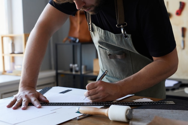 Abgeschnittene Aufnahme eines männlichen Handwerkers, der eine Schürze trägt, während er Muster im Kopierraum der Gerberwerkstatt zeichnet