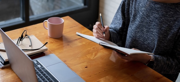 Abgeschnittene Fotohand einer Frau, die eine Liste schreibt und Notizen im Notizblock macht, die zu Hause mit dem Laptop online arbeitet oder lernt