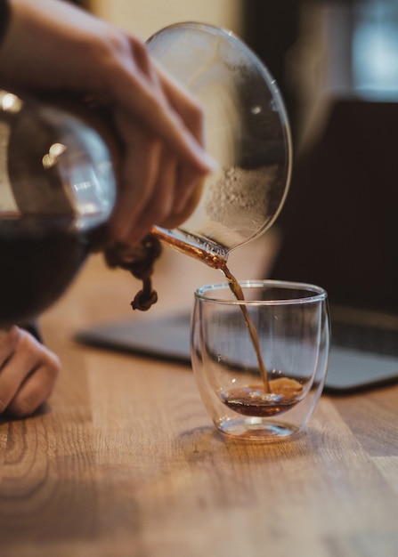 Abgeschnittene Hand, die Kaffee in ein Trinkglas auf einem Holztisch gießt