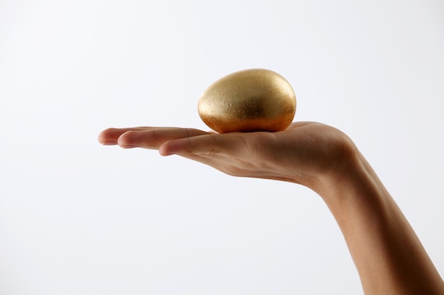 Abgeschnittene Hand einer Frau, die ein goldenes Ei vor weißem Hintergrund hält