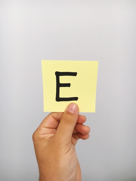 Foto abgeschnittene hand einer person, die den buchstaben e auf einer klebenden notiz gegen grauen hintergrund hält