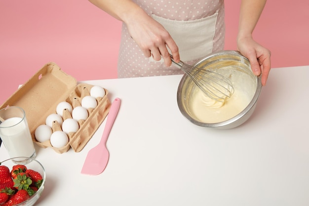 Abgeschnittene Köchin, Konditorin oder Bäckerin in Schürze, weißes T-Shirt, das Kuchen oder Cupcake am Tisch kocht, hält Schneebesenschüssel isoliert auf rosa Pastellhintergrund im Studio. Mock-up Copy Space Food-Konzept.