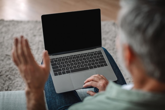 Abgeschnittener älterer kaukasischer Mann winkt mit der Hand und schaut auf Laptop mit leerem Bildschirm und unterhält sich im Innenraum