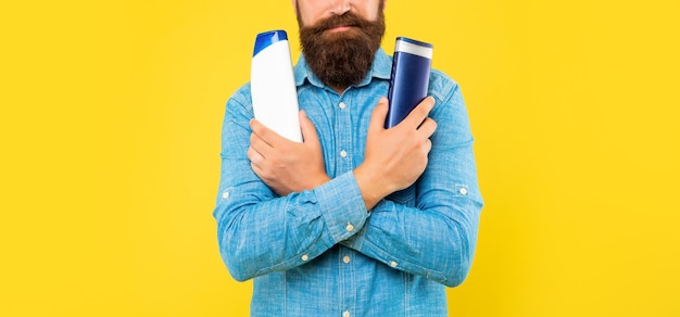 Abgeschnittener, brutaler, bärtiger Mann, der sich für eine Shampooflasche auf gelbem Hintergrund entscheidet und Toilettenartikel präsentiert