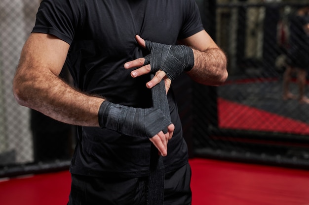 Abgeschnittener Mann in schwarzer Sportkleidung, der sich auf einen harten Kampf vorbereitet und die Faust in Sportschutzbandagen einwickelt. auf dem Ring stehen und sich auf den Mma-Kampf vorbereiten