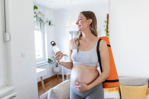 Abgeschnittener Mittelteil einer schwangeren Frau, die eine Yogamatte und eine wiederverwendbare Wasserflasche hält