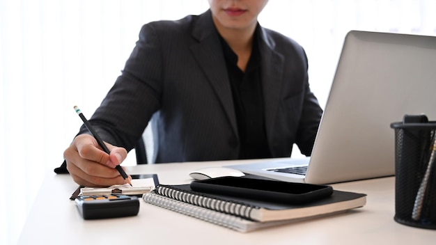 Abgeschnittener Schuss Geschäftsmann schreibt Informationen auf Notebook und benutzt Laptop im Büro