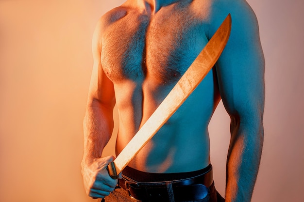 Abgeschnittenes Bild des Mannes mit scharfer Machete in der Hand. Sexueller muskulöser Kerl mit nacktem sportlichem Oberkörper. Getrennt auf beige Hintergrund mit blauem Licht. Studioshooting