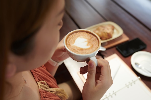 Abgeschnittenes Bild einer Frau, die im Café eine Tasse Cappuccino trinkt und Pläne für den Tag schreibt