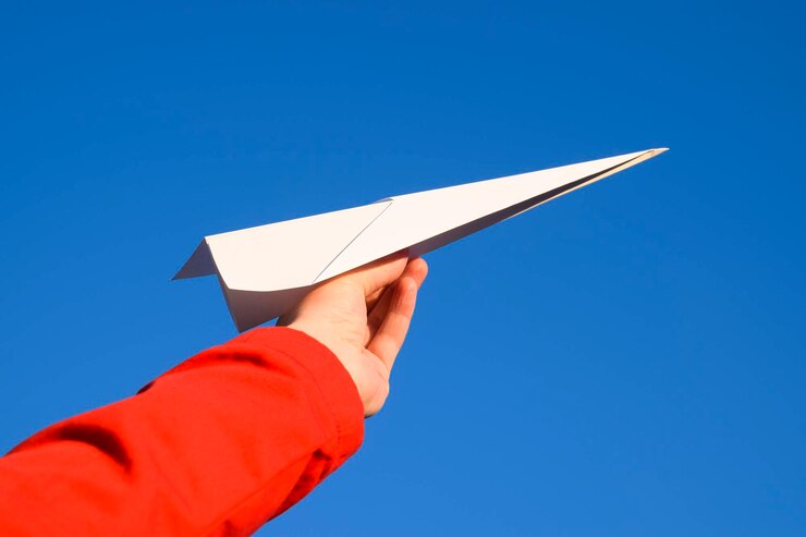 Foto abgeschnittenes bild einer hand, die ein papierflugzeug gegen einen klaren blauen himmel hält