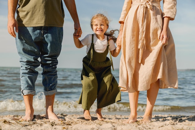 Abgeschnittenes Bild einer jungen Familie, die mit einem glücklichen süßen Mädchen am Strand steht