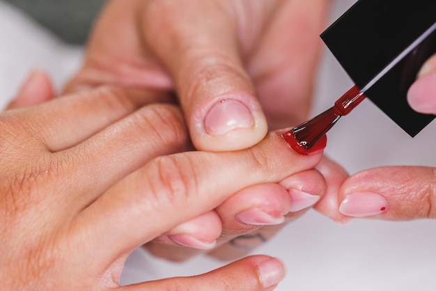 Abgeschnittenes Bild einer nicht erkennbaren Kosmetikerin, die einer Frau während einer Manikürebehandlung in einem Schönheitssalon roten Nagellack aufträgt.