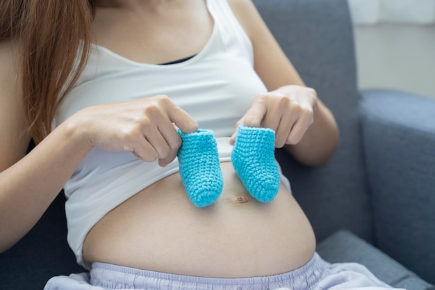 Abgeschnittenes Bild einer schönen schwangeren jungen Frau, die ein Paar süße Babysocken auf ihrem nackten Bauch hält und zu Hause auf ihrem Sofa liegt