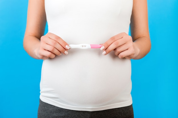 Abgeschnittenes Bild einer schwangeren Frau in Heimkleidung, die einen positiven Schwangerschaftstest mit zwei Streifen auf blauem Hintergrund zeigt. Gesunde Schwangerschaft. Platz kopieren.