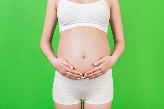 Abgeschnittenes Bild einer schwangeren Frau in weißer Unterwäsche, die ihren Bauch auf grünem Hintergrund hält. Zukünftige Mutter wartet auf ihr Baby. Platz kopieren.