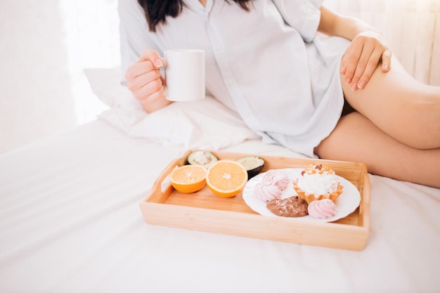 Abgeschnittenes Bild einer sexy attraktiven Frau in Unterwäsche mit nackten Beinen, die eine Tasse Kaffee hält und ein gesundes, leckeres Frühstück auf einem Holztablett im Bett im Hotelzimmer hat. Platz kopieren.