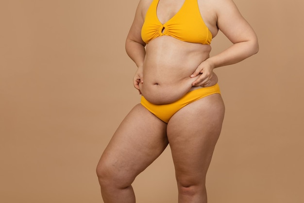 Abgeschnittenes Bild einer übergewichtigen Frau, die sich in einer gelben Hose, die den Bauch herunterbaumelt, einen fetten, nackten, großen, übermäßigen Bauch versteckt