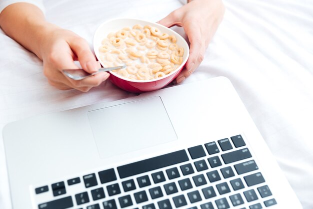 Abgeschnittenes Bild einer weiblichen Hände mit Schüssel Müsli und Laptop-Computer