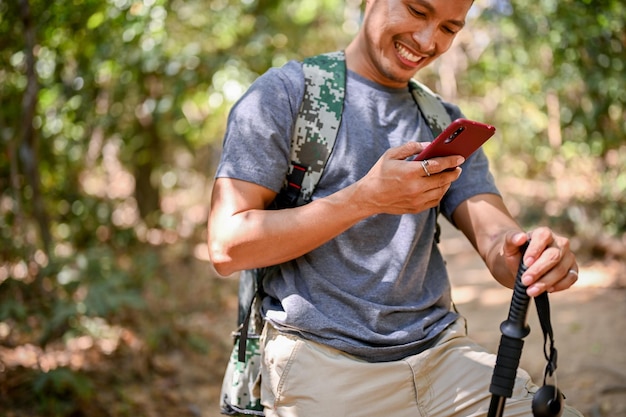 Abgeschnittenes Bild eines glücklichen asiatischen männlichen Reisenden mit Trekkingausrüstung mit seinem Smartphone
