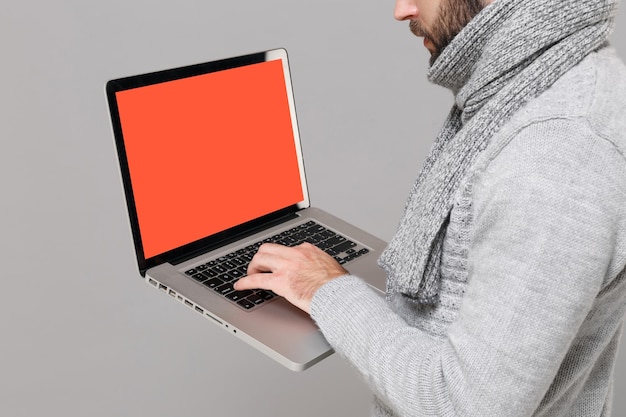 Abgeschnittenes Bild eines jungen Mannes in grauem Pullover, Schal isoliert auf grauem Hintergrund. Beratung zur Online-Gesundheitsbehandlung in der kalten Jahreszeit. Mock-up-Kopienbereich. Halten Sie einen Laptop-Computer mit leerem, leerem Bildschirm.