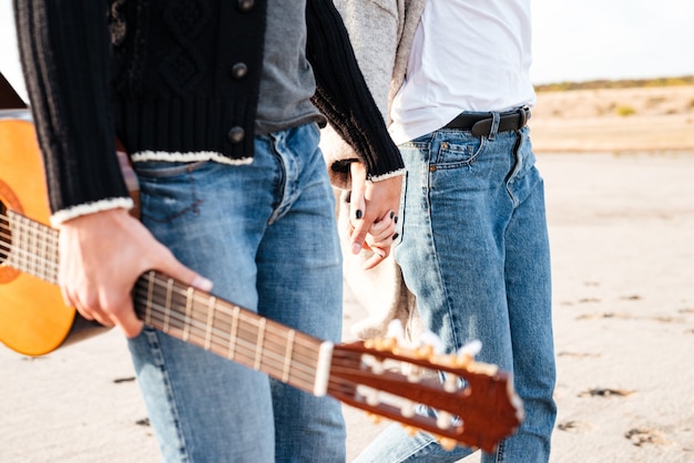 Abgeschnittenes Bild eines jungen zufälligen Paares, das mit Gitarre geht und Händchen hält am Strand