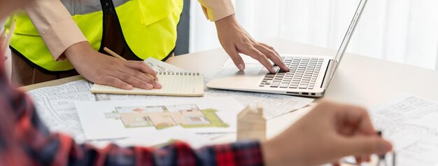 Foto abgeschnittenes bild eines professionellen ingenieurenteams, das an einer blaupause arbeitet, während sein mitarbeiter an einem laptop am tagungstisch arbeitet, wobei eine blaupause und ein holzblock herum verstreut sind.