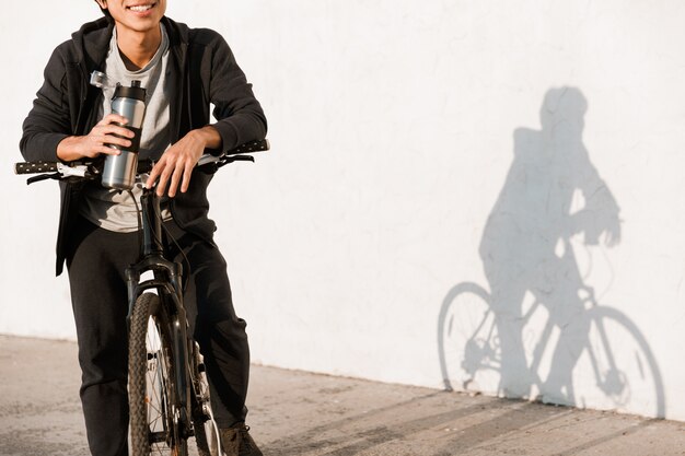 Abgeschnittenes Bild lächelnder asiatischer Mann, der im Freien Fahrrad fährt und Wasser aus einer Flasche trinkt