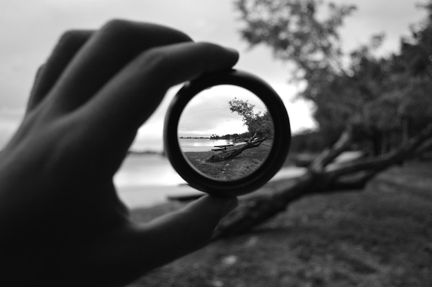 Foto abgeschnittenes bild von einer hand, die eine linse gegen bäume am seeufer hält