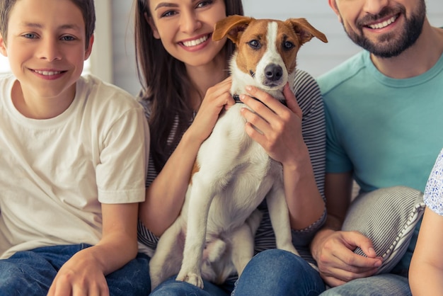 Abgeschnittenes Bild von schönen jungen Eltern und ihrem Sohn, die in die Kamera schauen und lächeln, mit ihrem süßen Hund zu Hause auf dem Sofa sitzen