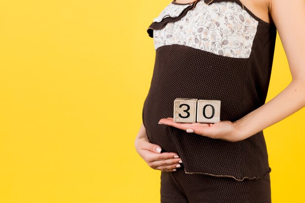 Abgeschnittenes Bild von Würfeln mit dreißig Wochen Schwangerschaft in den Händen der schwangeren Frau, die braune Pyjamas auf gelbem Hintergrund tragen. Anstehende Lieferung. Platz kopieren.