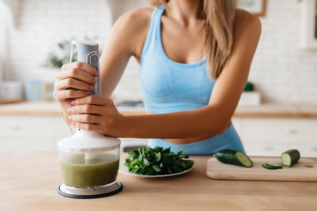 Abgeschnittenes Foto einer Dame in blauem Oberteil, die einen Küchenmixer für die Zubereitung eines gesunden Gemüsesmoothie verwendet