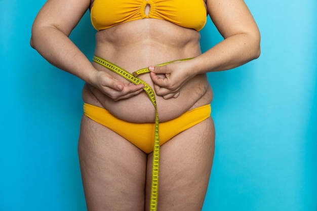 Abgeschnittenes Foto einer dicken Frau, die in gelben Badehosen steht und überschüssigen nackten Bauch zeigt, der die Taille mit Klebeband misst