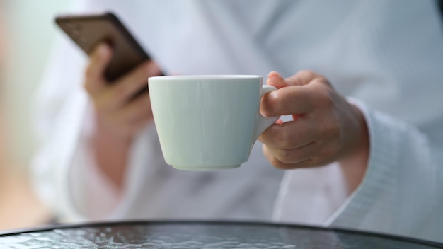 Abgeschnittenes Foto einer Frau in einem Frottee-Bademantel mit Handy und Tasse