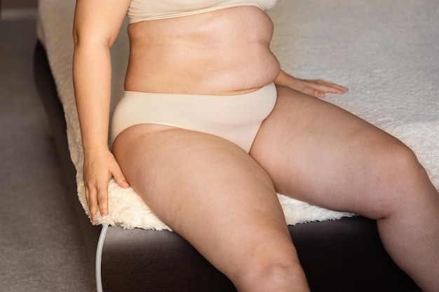Abgeschnittenes Foto einer prallen übergewichtigen Frau, die sich in nackten BH-Unterhosen auf ein beiges Plaid stützt und einen übermäßig nackten Bauch zeigt