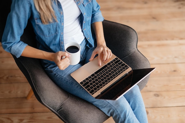 Abgeschnittenes Foto einer unbekannten Dame, die eine Kaffeetasse hält und ihren Laptop benutzt, während sie in einem Sessel sitzt